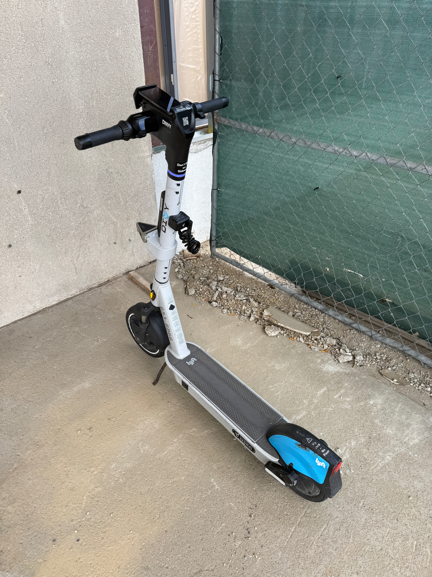 A white electric scooter is parked on a concrete surface next to a chain-link fence.
